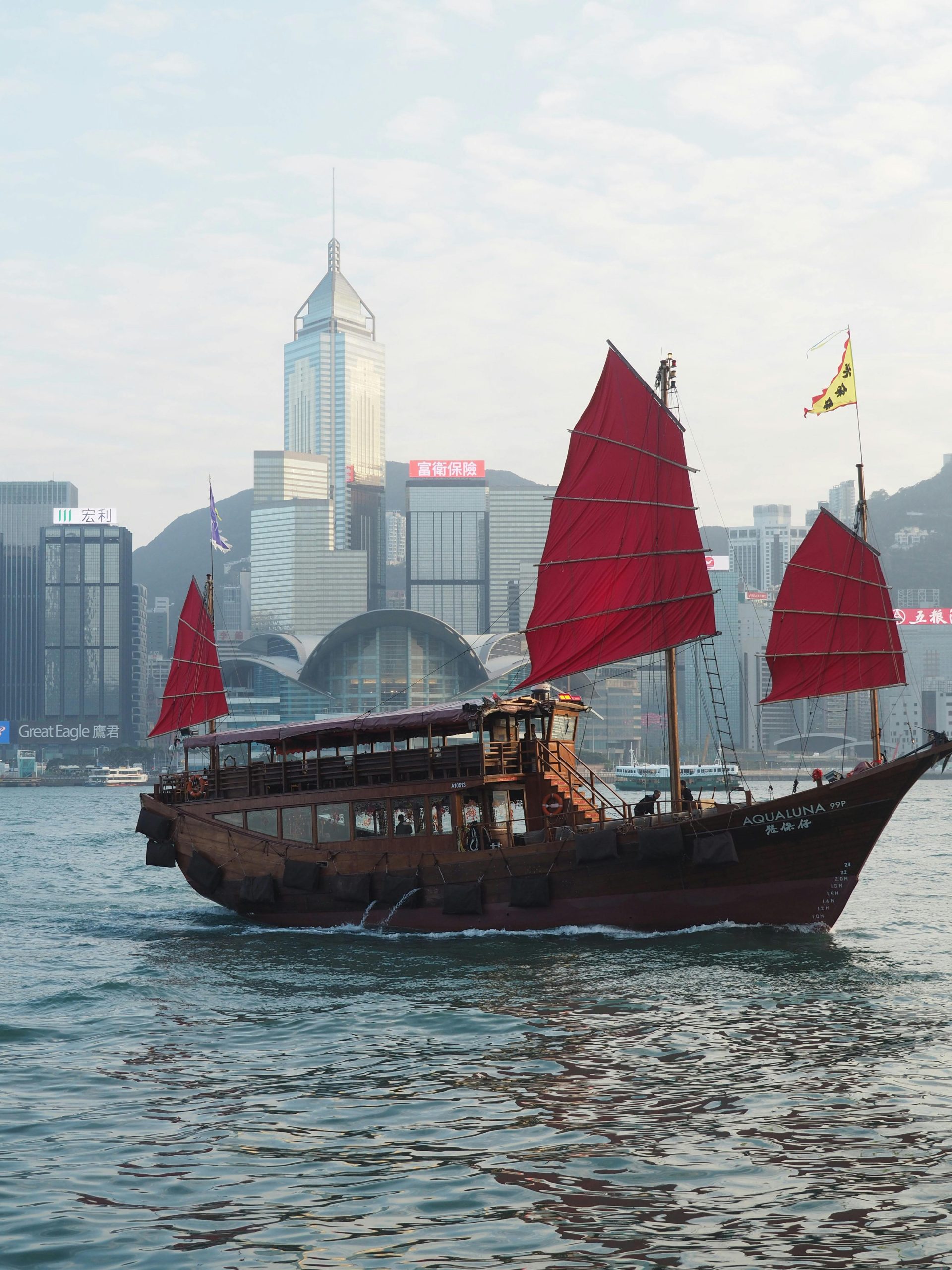 Iconic Hong Kong junk boat with red sails cruising in Victoria Harbor against a skyline backdrop.
